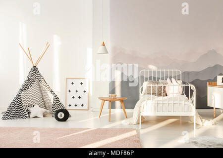 Patterned teepee standing in bright kids room interior with white bed, mountain wallpaper and powder pink carpet Stock Photo