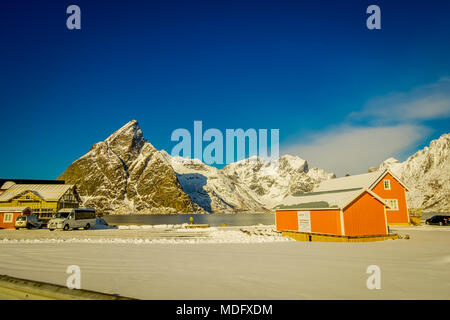 Henningsvaer, Norway - April 04, 2018: Outdoor view of wooden houses of a typical little village close to the shore at lake with a huge mountain behind covered with snow in Lofoten Islands Stock Photo
