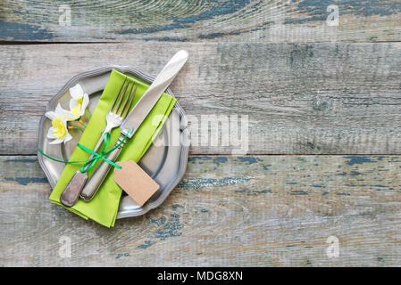 Ancient metal plate with a green napkin, vintage cutlery and spring flowers on the background of an old wooden table with space for text Stock Photo