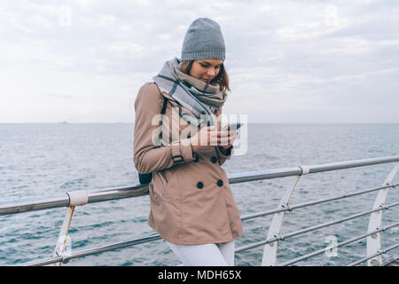 Smiling young woman looks down on smart phone on the seafront. Cute stylish girl stands leaning on the embankment railing at cloudy spring day, sea at Stock Photo