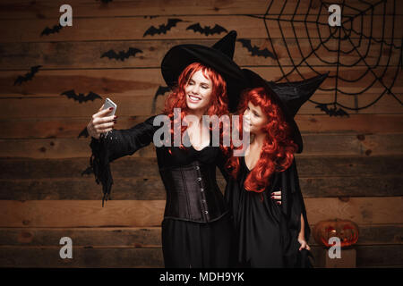 Halloween Concept - Beautiful caucasian mother and her daughter with long red hair in witch costumes taking a selfie with smartphone  ready for celebrating Halloween. Stock Photo