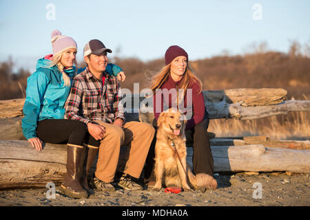 A young couple and a friend with a dog sit on a piece of driftwood on a beach looking out to the ocean at sunset Stock Photo