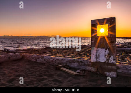 A sunburst shines bright through the window of a weathered, wooden door propped up on Iona beach; Richmond, British Columbia, Canada Stock Photo