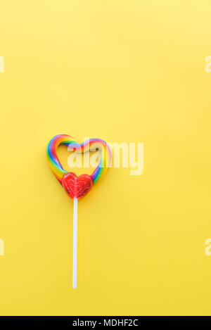 Cute heart-shaped rainbow swirl lollipop on solid yellow background portrait format Stock Photo