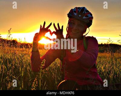 girl in a bicycle helmet on the field shows the symbol of the heart. Hands of woman showing heart shape with sunset in the background Stock Photo