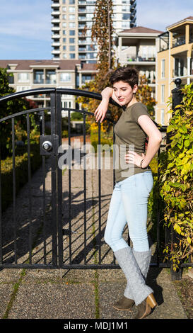 A young woman stands leaning against a metal gate looking confident with housing in the background; New Westminster, British Columbia, Canada Stock Photo