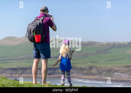 A man and girl child standing on a clifftop looking out to sea in summer clothes. He is taking a photo with a mobile phone. Stock Photo