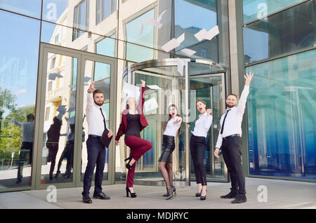 Office workers throw paper up against the background of a multi-storey glass office building. Group of young people. Five are happy about the successf Stock Photo