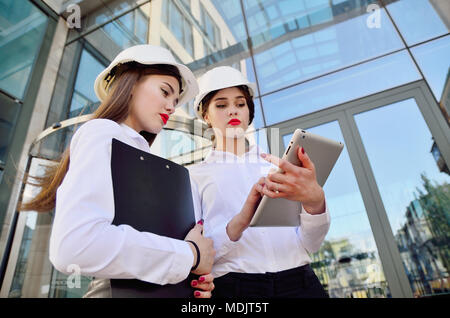 Girls in building white helmets with tablets in hands on the background of a glass office building. Women are engineers. Female professions. Stock Photo