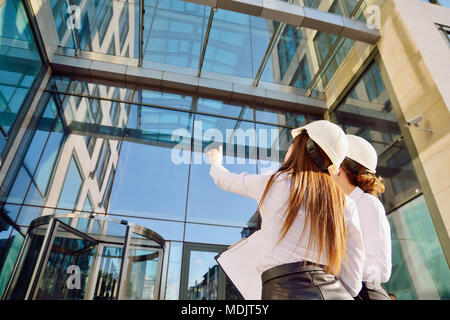 Girls in building white helmets with tablets in hands on the background of a glass office building. Women are engineers. Female professions. Stock Photo