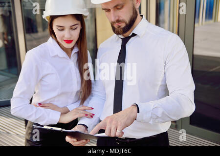 A girl with a man in construction white helmets and white shirts with drawings in hands against a glass office building. Engineers. Planning of constr Stock Photo