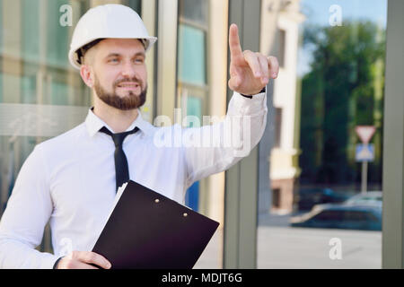 A man working in a white helmet and white shirt with a tablet in his hands clicks his index finger in the virtual screen. Engineer. Planning of constr Stock Photo