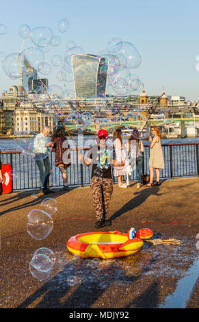 London, UK, 19th April, 2018. An entertainer on the south bank of the Embankment of the River Thames at Bankside blows bubbles to entertain children against a skyline background of iconic modern skyscraper buildings in the City of London. The sunny fine weather on the warmest April day in decades brought out good-natured crowds to enjoy the sunshine. Credit: Graham Prentice/Alamy Live News. Stock Photo