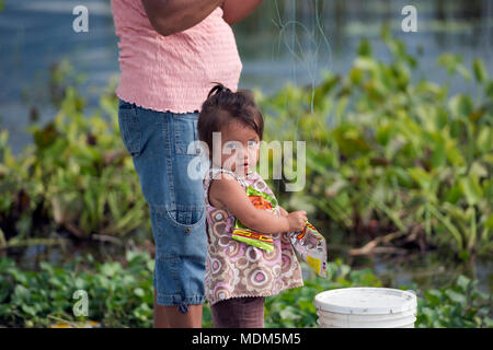 A young, indigenous Itza Maya girl eats candy while her mother fishes in Lake Peten, northern Guatemala. Stock Photo