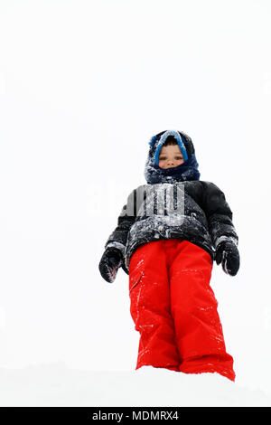 Looking up at a young boy (5 yr old) dressed in outdoor winter clothes. He is stood on top of a pile of snow with an almost white sky behind.