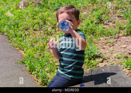 Thirsty little boy sitting on a wooden bench and drinking water. Stock Photo