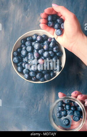 Woman putting freshly gathered blueberries from a jar into a small bowl. Table painted in blue in the background Stock Photo