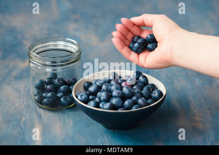 Woman putting freshly gathered blueberries from a jar into a small bowl. Table painted in blue in the background Stock Photo