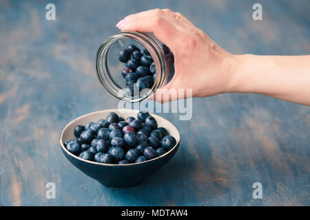 Woman putting freshly gathered blueberries from a jar into a small bowl. Table painted in blue in the background Stock Photo