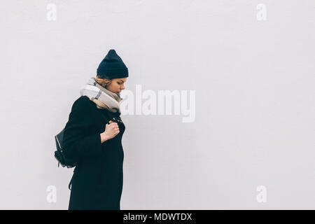 Side view of hipster young woman standing outdoors in cold weather on grey wall background. Girl wearing coat, hat and scarf holding a backpack, copy  Stock Photo