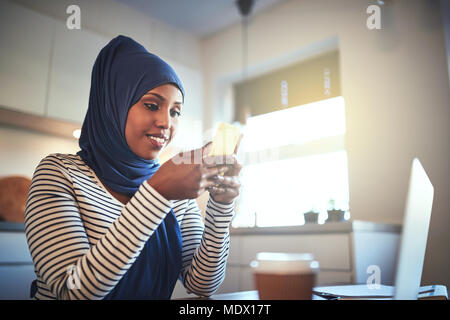 Smiling young Arabic woman wearing a hijab sitting in her kitchen at home sending a text message and using a laptop Stock Photo