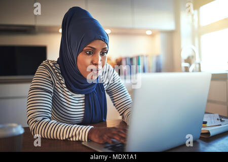 Young Arabic woman wearing a hijab sitting at her kitchen table using a laptop while working from home Stock Photo
