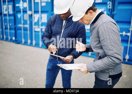 Two engineers comparing inventory lists on a clipboard and a digital tablet while standing by freight containers on a large commercial shipping dock Stock Photo