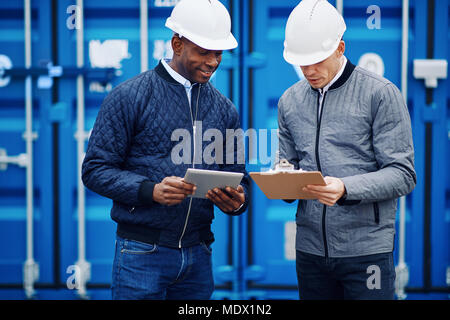 Two smiling engineers standing by freight containers on a large commercial shipping dock comparing inventory lists on a clipboard and a tablet Stock Photo