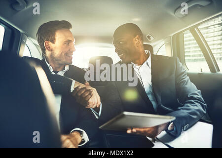 Two smiling businessman shaking hands together while sitting in the backseat of a car driving through the city Stock Photo