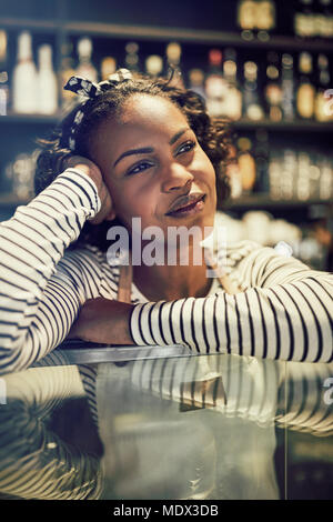 Smiling young African woman leaning on the counter of her trendy cafe looking deep in thought Stock Photo