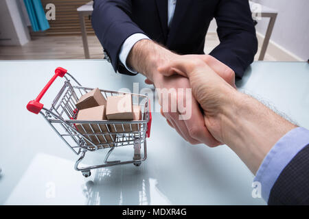 Close-up Of Two Businesspeople Shaking Hands Near Shopping Cart Full Of Cardboard Boxes Stock Photo