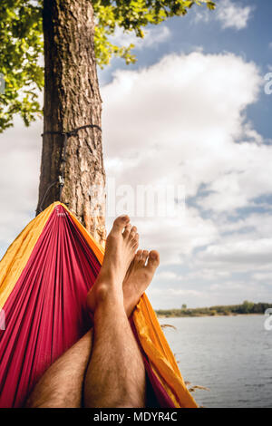 Relaxing in the hammock at the beach under a tree, summer day. Barefoot man laying in hammock, looking on a lake, inspiring landscape Stock Photo