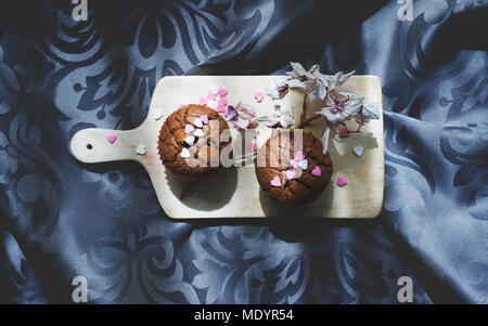 Still Life with Two Chocolate Muffins on Wooden Cutting Board with Sprinkling Hearts and Dry Orchid on Silver Ornamental Background. Stock Photo