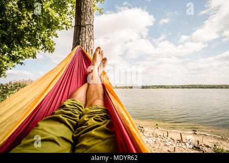 Relaxing in the hammock at the beach under a tree, summer day. Barefoot man laying in hammock, looking on a lake, inspiring landscape Stock Photo
