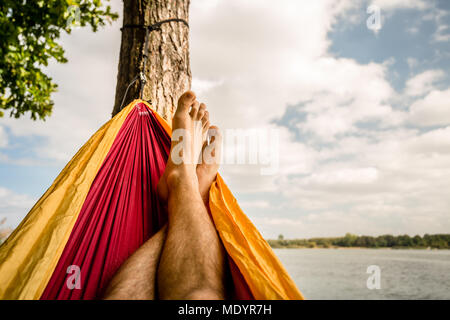 Relaxing in the hammock at the beach under a tree, summer day. Barefoot man laying in hammock, looking on a lake, inspiring landscape Stock Photo