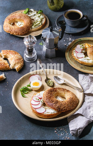Variety of homemade bagels with sesame seeds, cream cheese, pesto sauce, eggs, radish, herbs served on ceramic plate with ingredients and coffee above Stock Photo