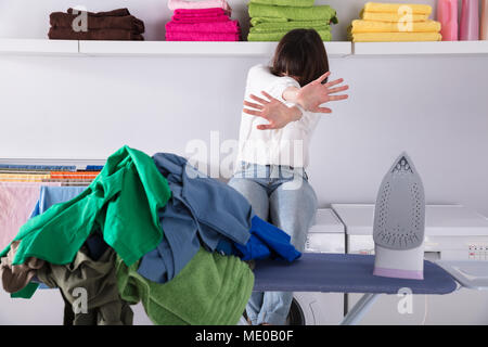 Upset Woman Sitting Behind Pile Of Clothes And Iron On Ironing Board Stock Photo