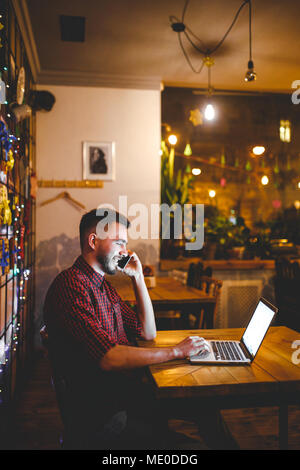 Young handsome Caucasian man with beard and toothy smile in red shirt works behind laptop, hands on keyboard sitting at wooden table. Uses calls on mo Stock Photo