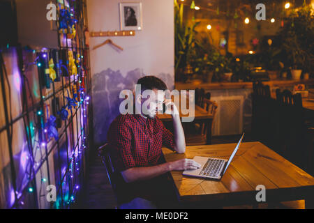 Young handsome Caucasian man with beard and toothy smile in red shirt works behind laptop, hands on keyboard sitting at wooden table. Uses calls on mo Stock Photo