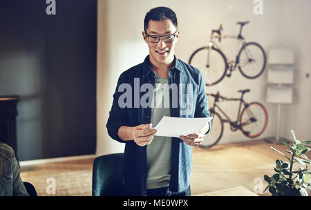 Smiling Asian man wearing glasses standing at a table discussing paperwork with colleagues in a creative modern office Stock Photo