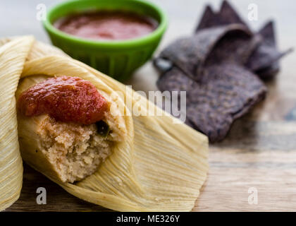 Partly unwrapped tamale served with blue corn tortillas and tomato sauce on wooden table side view - Mexican Food Stock Photo