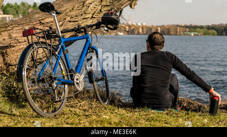 A peaceful atmosphere near the water with a man resting in nature close to his bicycle. The bike has a helmet on it and the cyclist is holding a water Stock Photo
