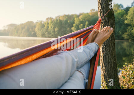 Personal perspective of woman relaxing on hammock by the lake in Springtime season, barefoot, sunlight reflection on lake surface. Travel laziness Stock Photo