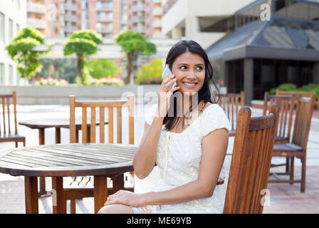 Closeup portrait, young, happy beautiful woman in white dress sitting down outside at brown table, speaking on cell phone, isolated background of buil Stock Photo