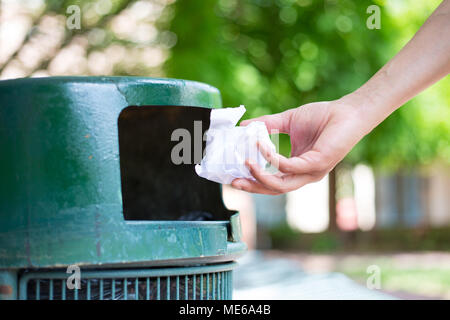 Closeup cropped portrait of someone tossing crumpled piece of paper in trash can, isolated outdoors green trees background Stock Photo