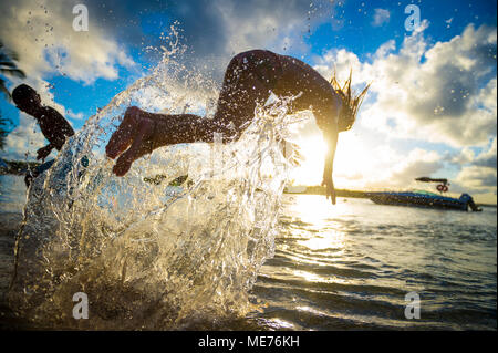 Silhoutte of unidentifiable young man jumping and splashing into a calm sea from the beach at sunset. Focus on splashing water. Stock Photo
