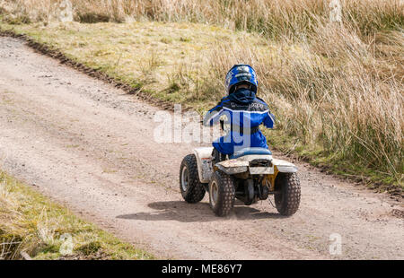 West Linton, Scottish Borders, Scotland, United Kingdom, April 21st 2018.  Spring sunshine in the countryside, with a young boy wearing a motorcycle helmet driving a small quad bike on a farm track Stock Photo