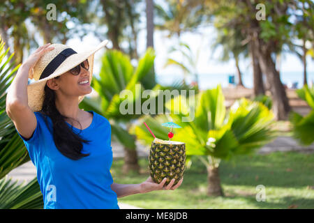 Closeup portrait, woman in blue shirt and brown straw hat holding pineapple mixed drink with rum at resort, isolated background of green palm trees Stock Photo