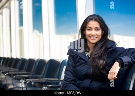 Closeup portrait of confident smiling happy pretty young woman sitting in winter fur coat waiting in airport terminal, isolated background black seats Stock Photo