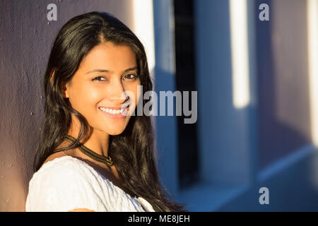 Closeup portrait of confident smiling happy pretty young woman in white dress during sunset, isolated background of shaded wall. Positive human emotio Stock Photo